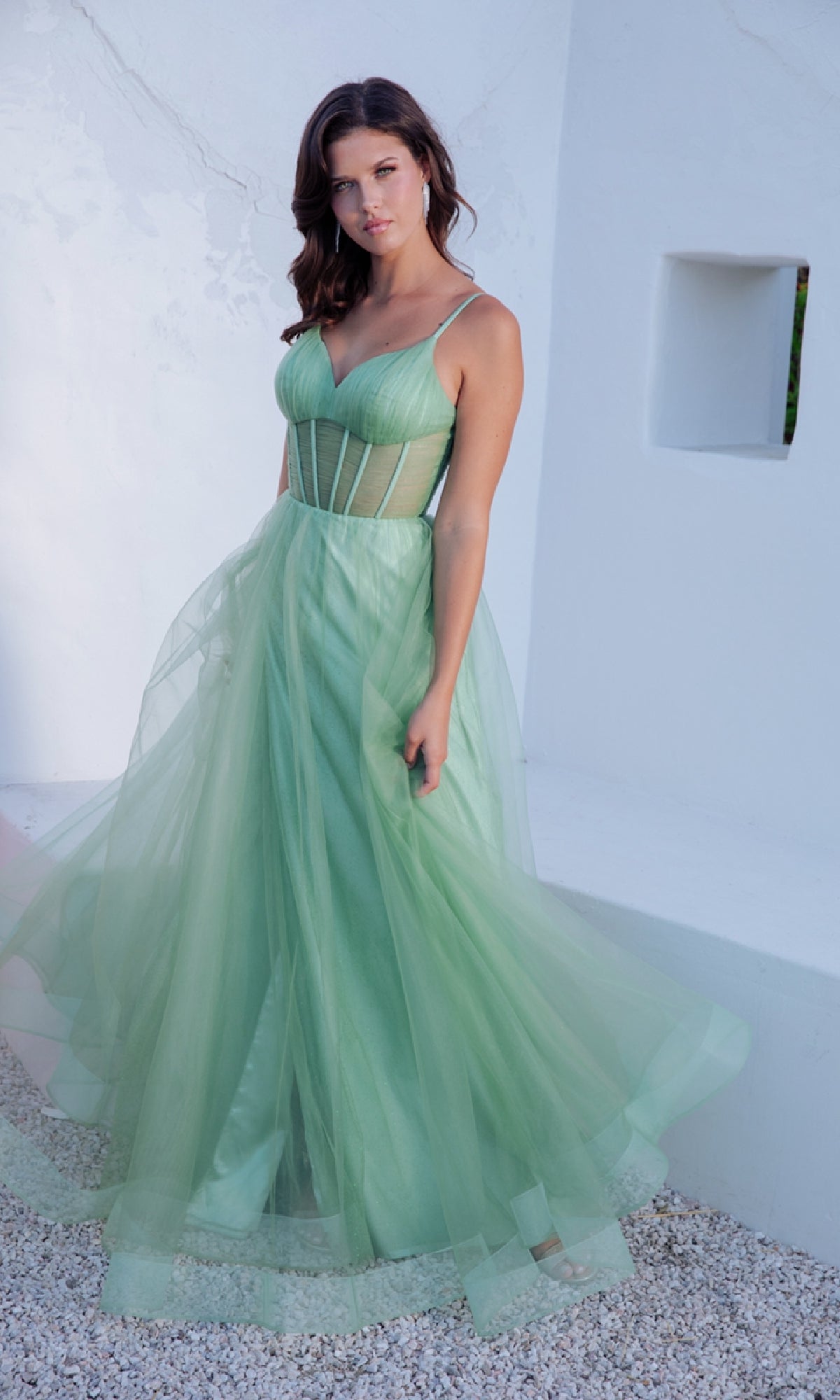 A woman in the Sheer-Corset Long Sage Prom Dress: Eureka 9199, featuring a fitted bodice and thin straps, stands against a white textured wall.